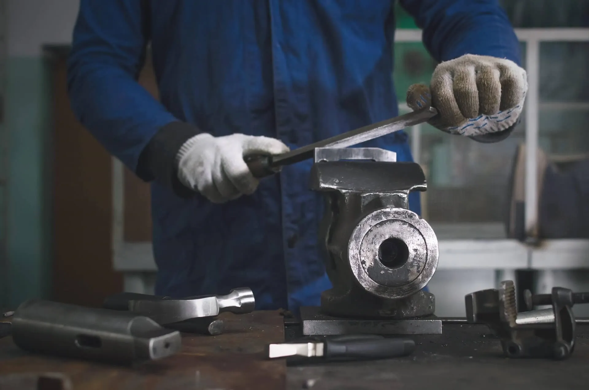 Man worker is working with iron detail on vise grip on blacksmith table and using a file.