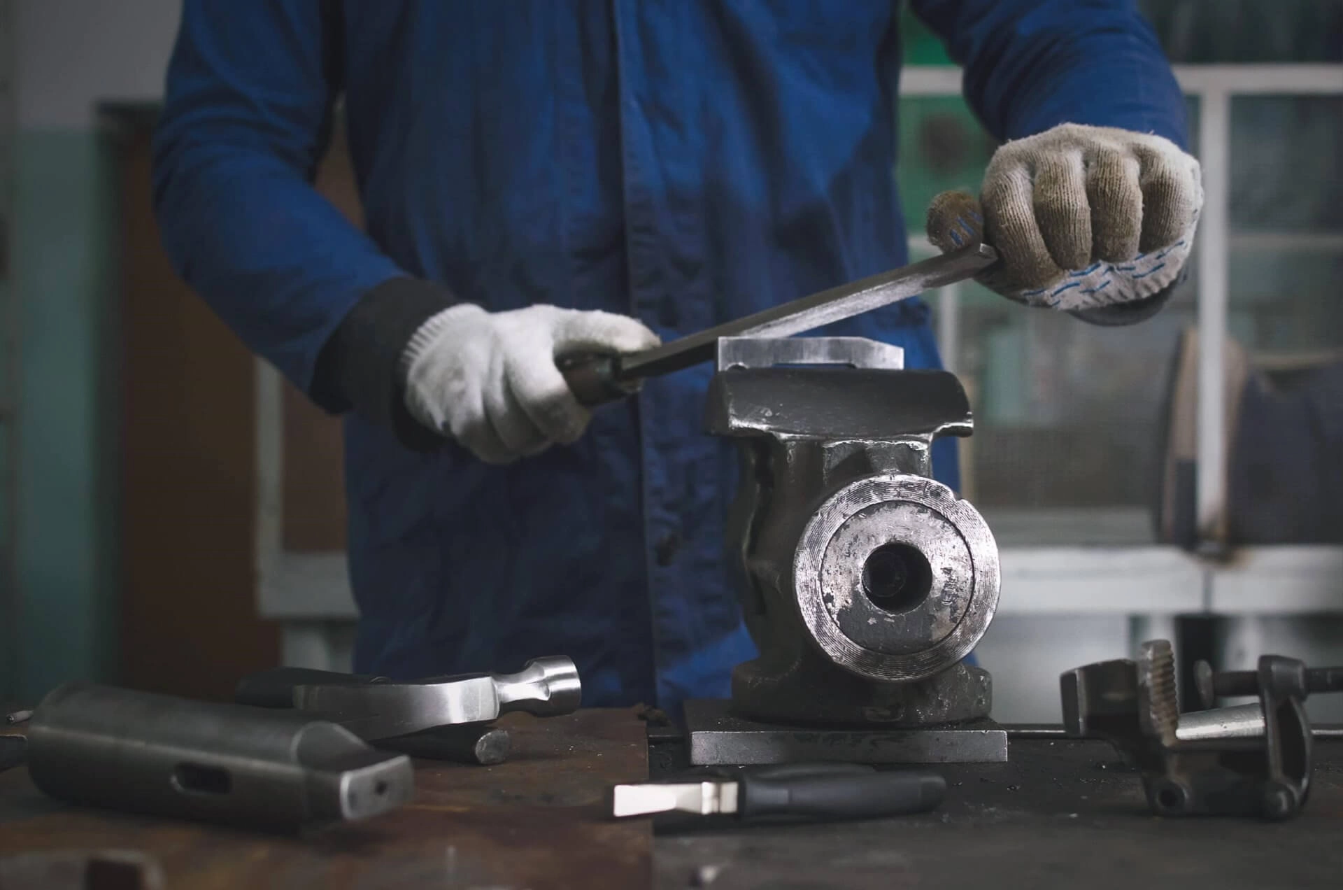 Man worker is working with iron detail on vise grip on blacksmith table and using a file.